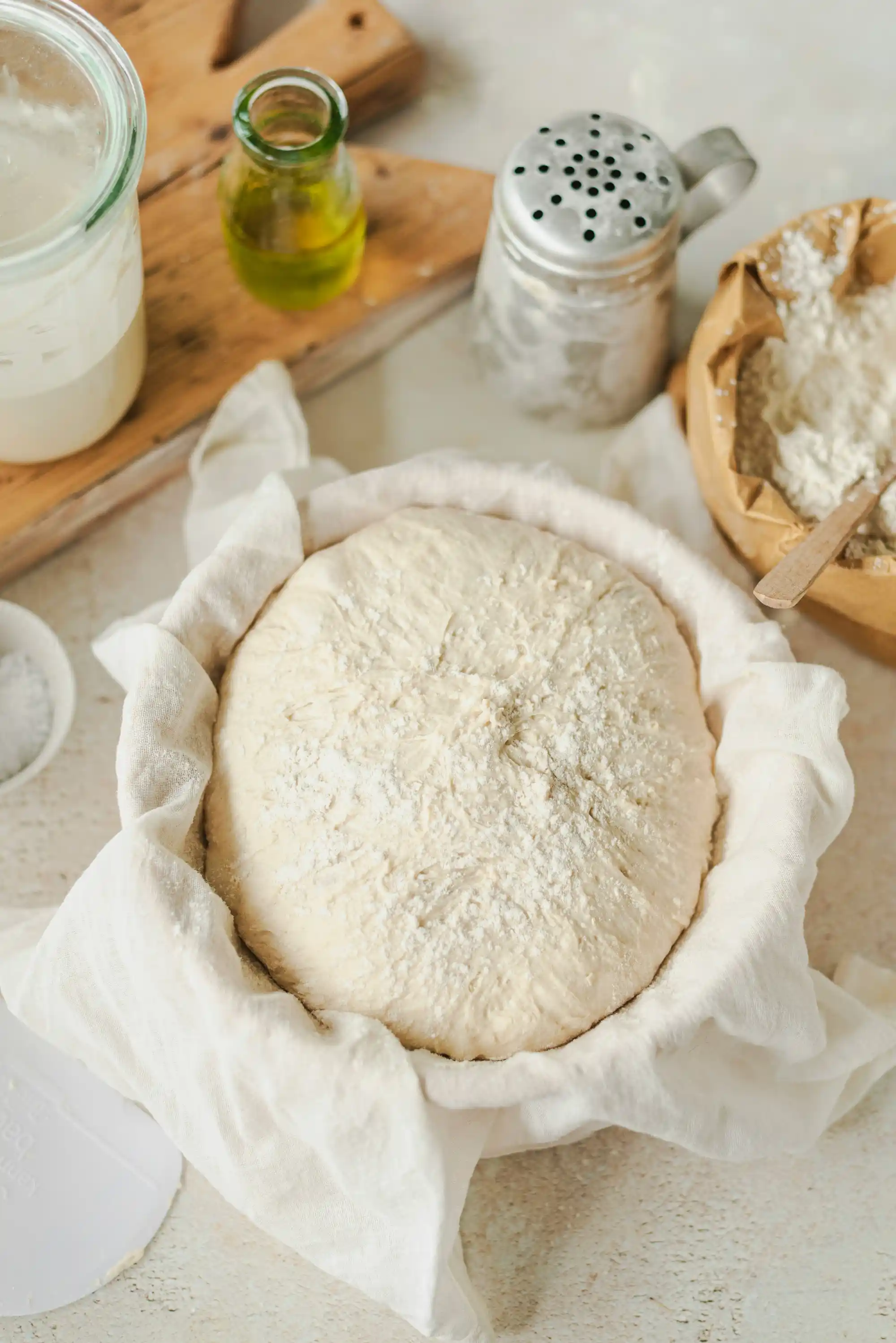 Пресен хлябов тесто, покрито с брашно, поставено в платнена кърпа върху дървена дъска. В близост има буркани с кисело мляко и масло, както и дървена купа, пълна с брашно, и метален сито за брашно. На заден план се вижда светло дървен плот.