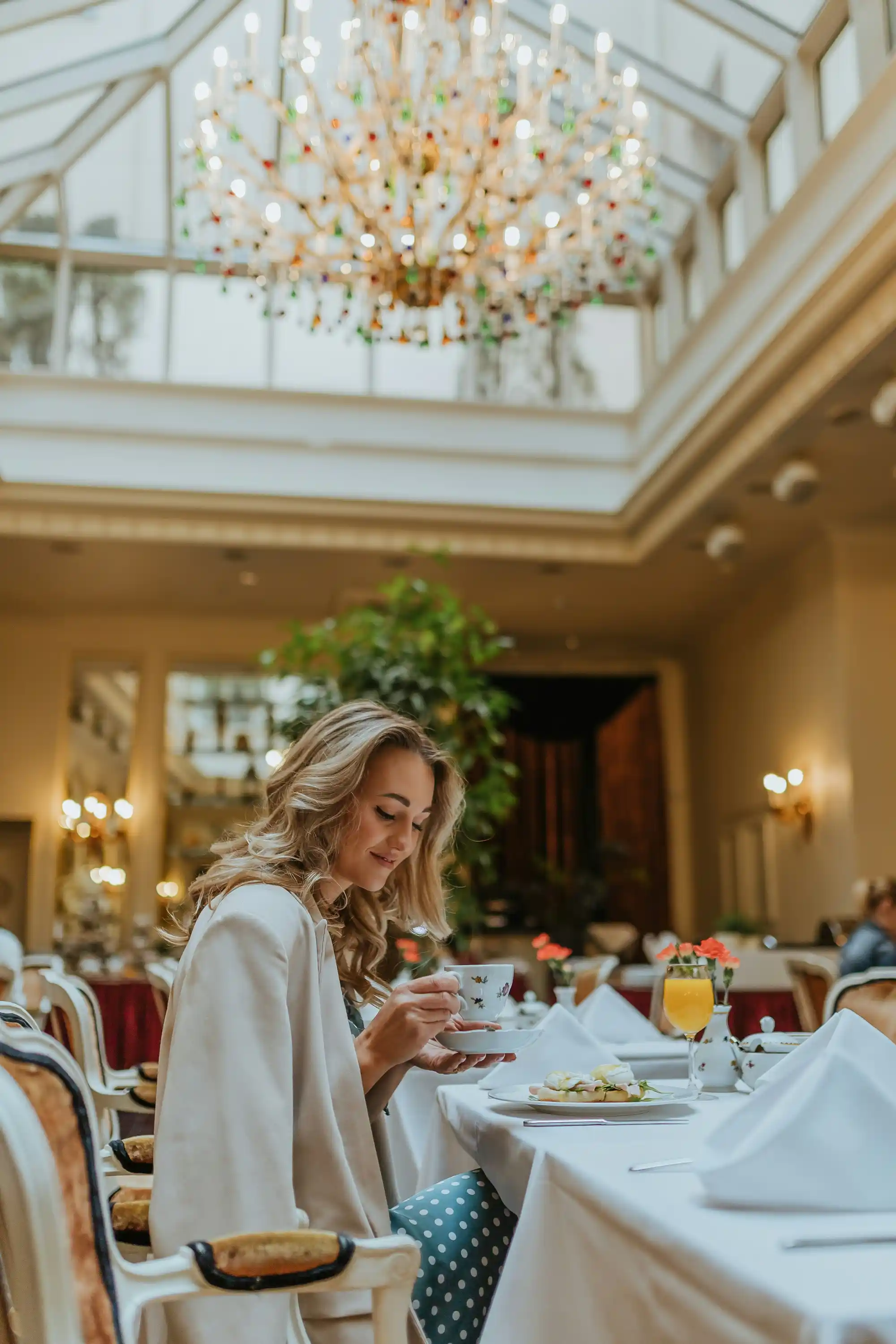 Млада жена седи на маса в елегантен ресторант, пие кафе и разглежда своя мобилно устройство. Над нея е окачен голям лустер с множество светещи кристали. Атмосферата е луксозна с високи тавани и богата растителност в задния план. Масите са подредени с бели покривки, а на масата пред жената има сервирани ястия и напитки.