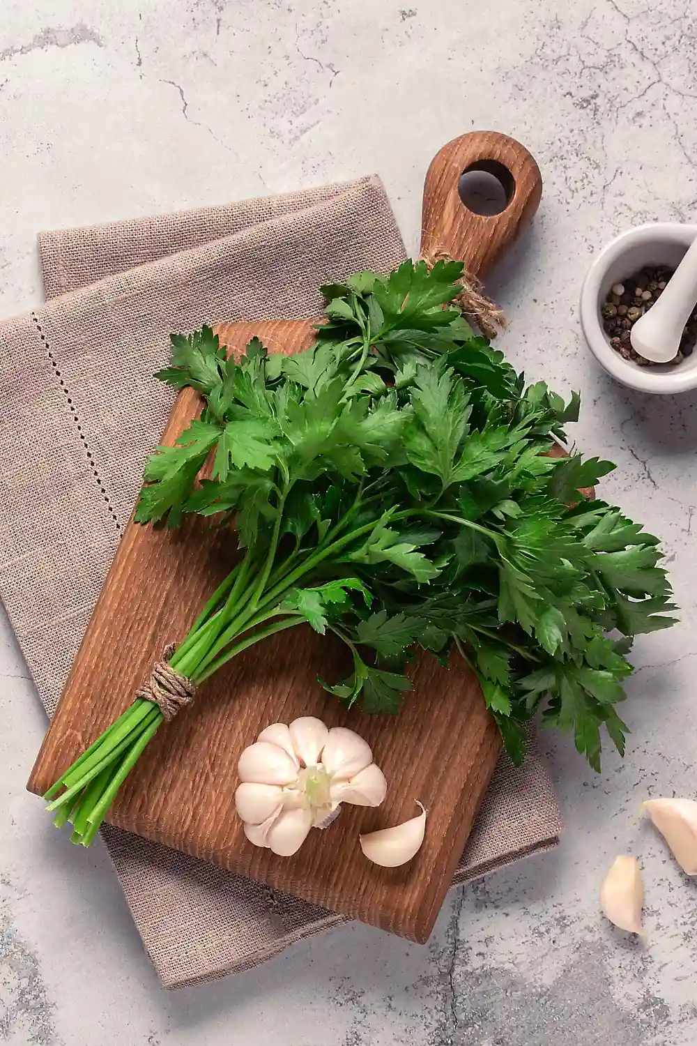 Вертикална снимка на дъска за рязане, поставена върху сива мраморна маса и кафява ленена салфетка. Върху дъската има връзка пресен магданоз и глава чесън с няколко отделени скилидки до нея. В десния горен ъгъл на снимката се вижда бяла керамична хаванче с пестил, пълна със смес от черни пиперки.