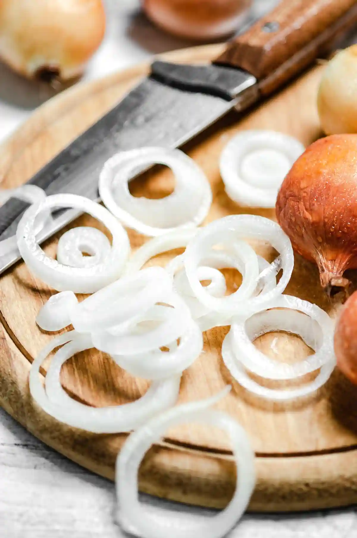 Дъска за рязане от дърво с нарязани на кръгчета луковици. На фона са видими още цели луковици и нож с черна дръжка.