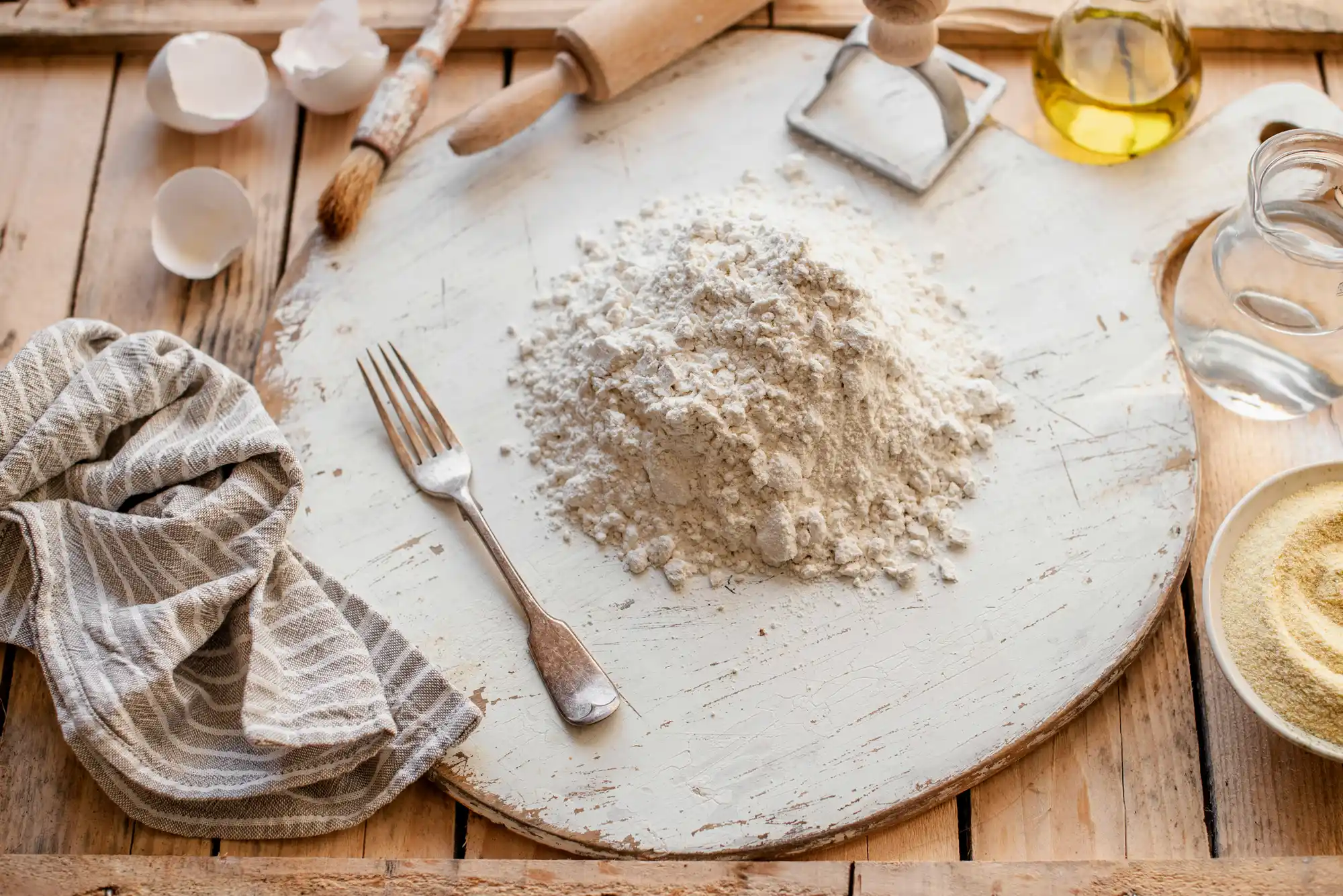 Кухненска сцена с разхвърляни съставки за готвене на дървена маса: купчина брашно върху бяла дъска, части от разбити яйца, вилица с брашно, дървена точилка, тъканена салфетка, контейнери с олио и вода и дървена четка.