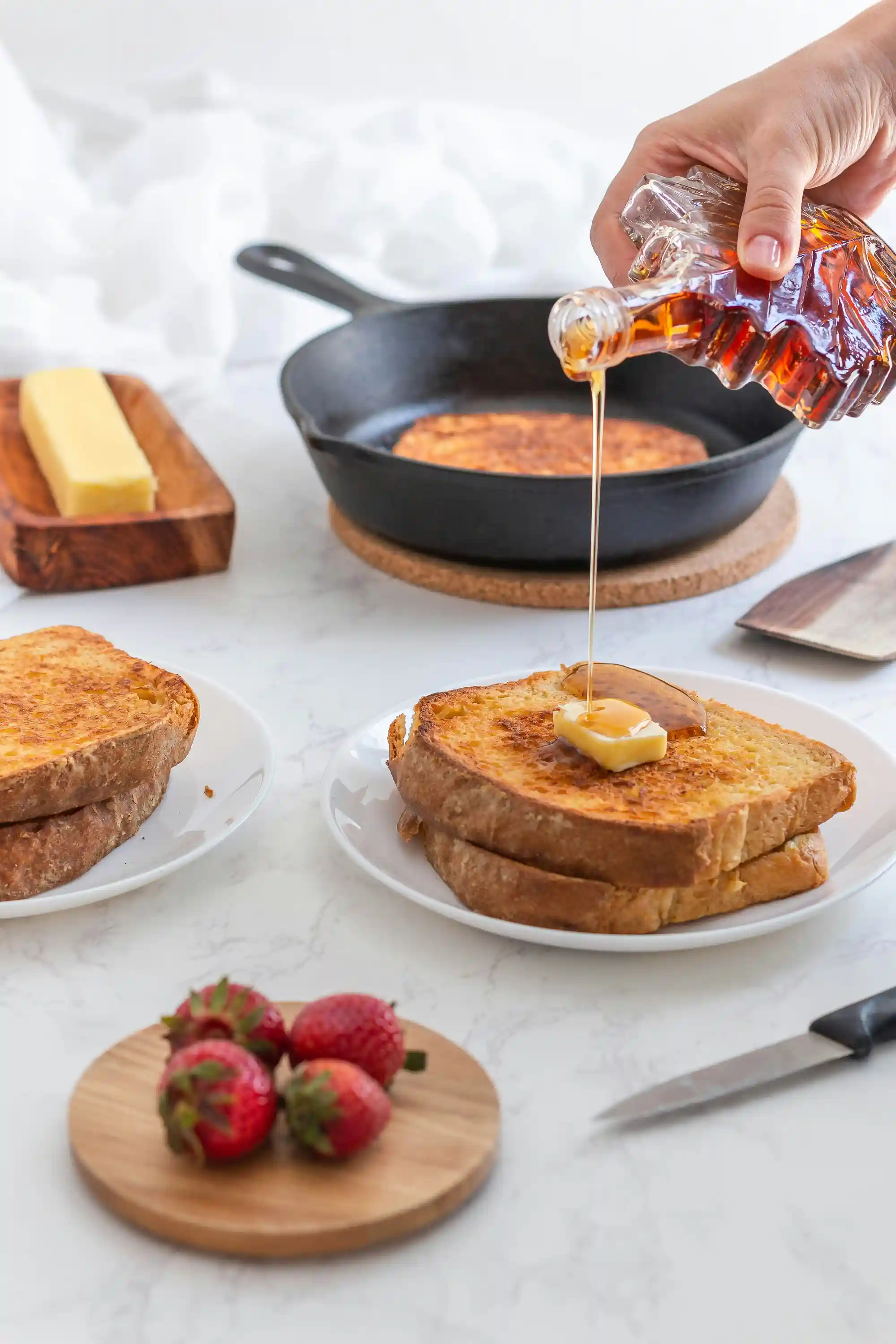 Човешка ръка изсипва мед от стъклена бутилка върху парче пържено френско тостче с кубче масло отгоре на бяла чиния, поставена върху бял мраморен плот. В близост има още две парчета тост на друга чиния и тенджера с тост на печка в заден план. Отстрани на дървена дъска са поставени и четири ягоди.