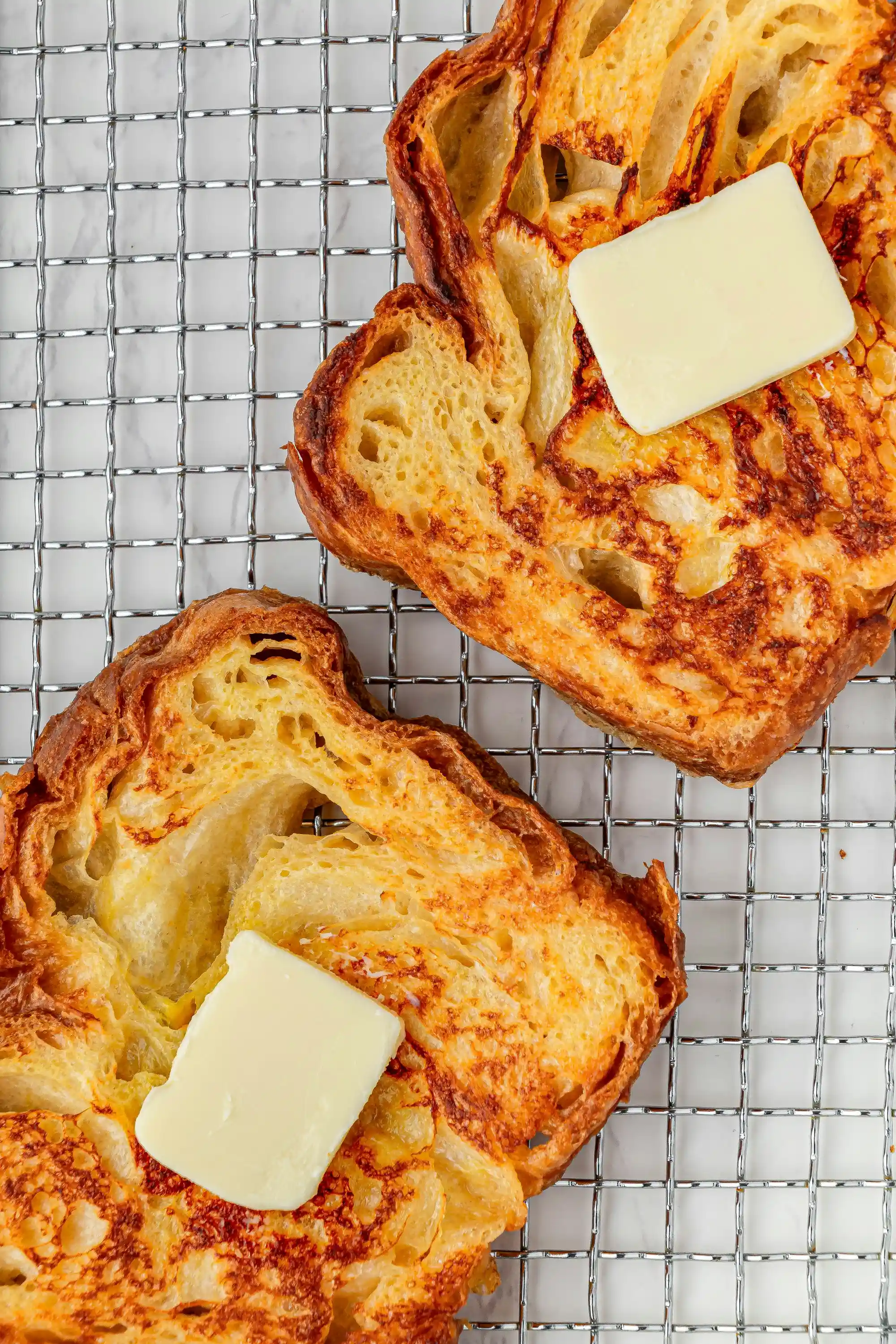 Две филии запечен хляб, поставени върху метална решетка, с парче масло върху всяка от тях. Хлябът има хрупкава златиста коричка и мека, пореста вътрешност, като маслото започва да се топи върху топлата повърхност.