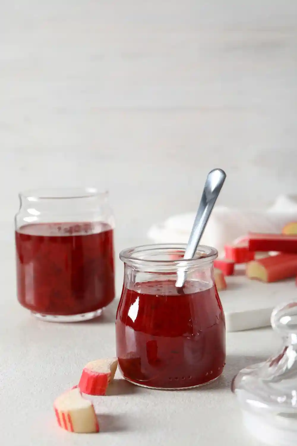 На снимката е изображен буркан с червен конфитюр и метална лъжица, поставен върху светла повърхност. До него са подредени още два буркана с конфитюр и нарязани на кръгчета парчета руло от ябълки. Фонът е светъл и нежен, като акцентира върху естетиката на храната.