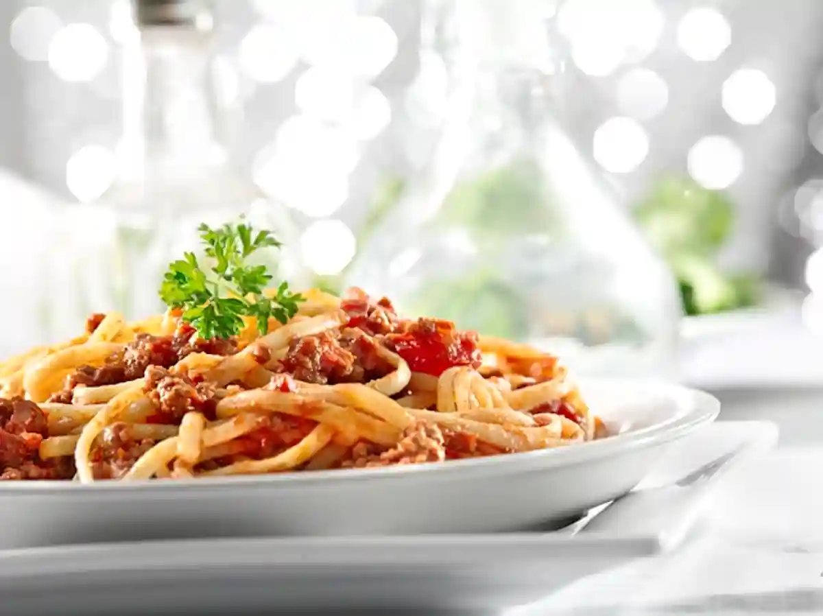 Чиния със спагети Болонезе, украсена с клонче магданоз, сервирана на бяла маса в светла стая, със замъглен фон, където се забелязват остро осветени контури и блясъци, предаващи усещане за топлина и приветливост.