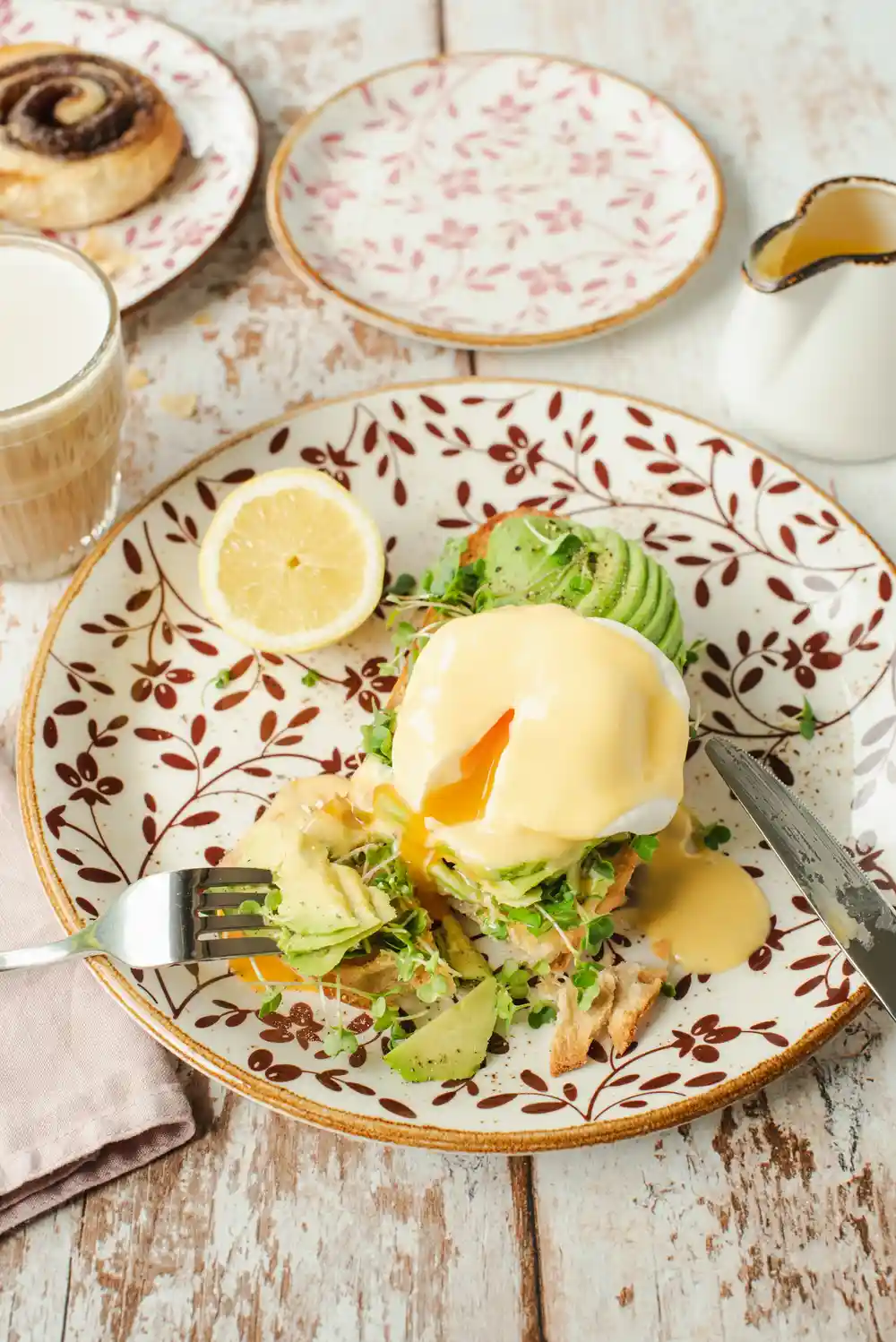 Поднесена на порцеланова чиния с декоративни шарки, тази изискана закуска включва ястие с варено яйце, покрито със холандез сос, сервирано върху слой от бързо задушени листа зелена салата и пухкава английска мафина, украсена с резени прясно авокадо. До мафината има парче лимон за допълнителна свежест. Чинията е част от по-голям сервиз за хранене, включително други чинии със същия декор и чаша с напиток, всичко разположено върху дървена маса с видими следи от употреба.