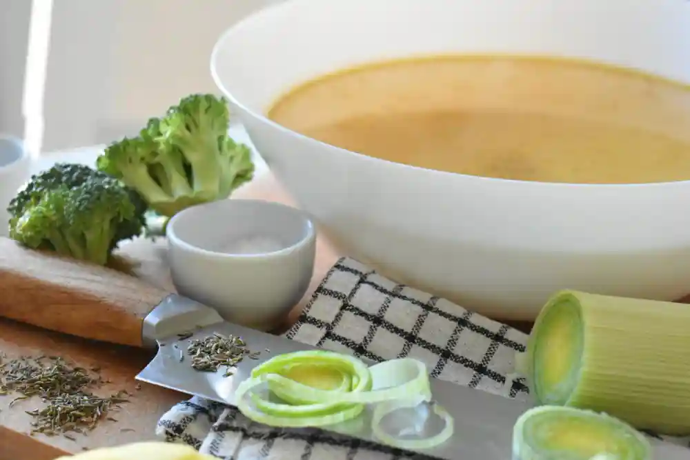 Снимка на кухненска сцена с голяма бяла купа супа от наситено жълт цвят, заобиколена от съставки за готвене - броколи, прясно нарязан праз, дървена дъска, кухненски нож и малка бяла купичка със сол. На преден план са разпръснати стръкове сушен чубриц.