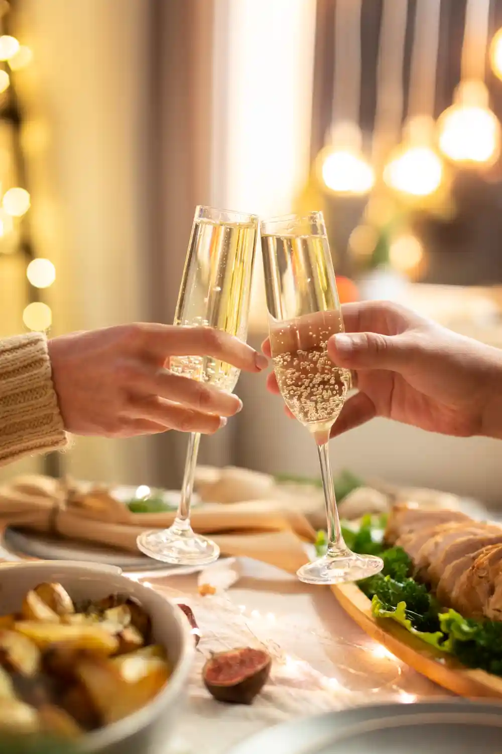 Двама души държат чаши с шампанско, чукват си в приятна обстановка с топла светлина и празнична украса. На масата има сервирани празнични ястия като пържено пиле и зеленчуци. Тази сцена излъчва атмосфера на празненство и уют.