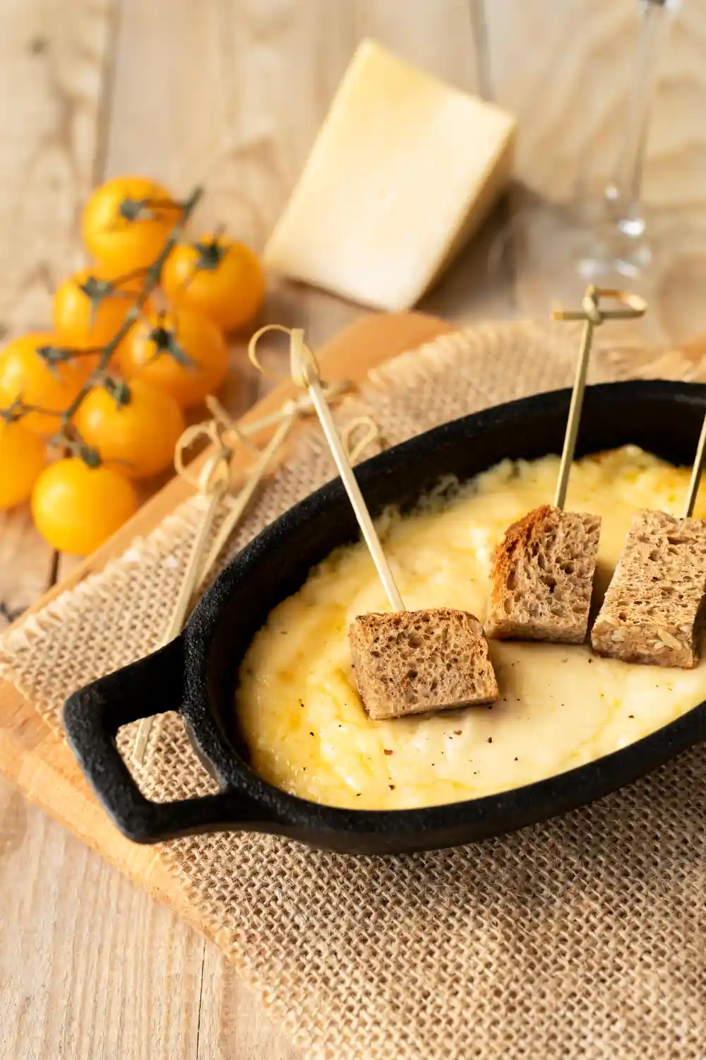 Порция расплавено сирене в черна чугунена тавичка, поднесена на дървена подложка с отпред избледнели домати на клонка и парче твърдо сирене в заден план. Над сиренето са забодени три парчета пълнозърнест хляб на клечки за зъби, а в десния горен ъгъл се вижда стъклена чаша.