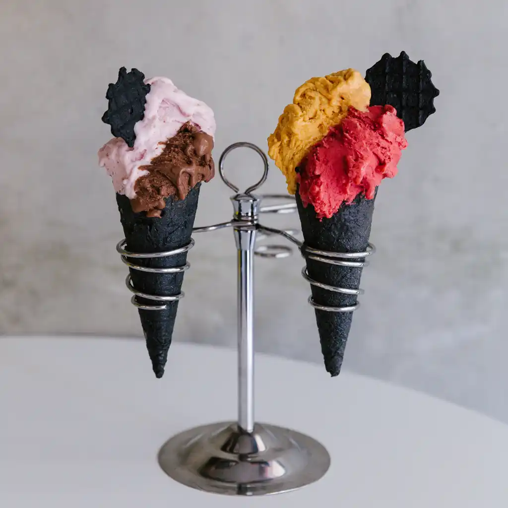 Два сладоледа в черни конуси, поставени в метална стойка с кръгла основа. Левият конус съдържа шоколадов и ягодов сладолед с черна декоративна фигурка, а десният - сладолед в оранжево и червено със същата такава фигурка.