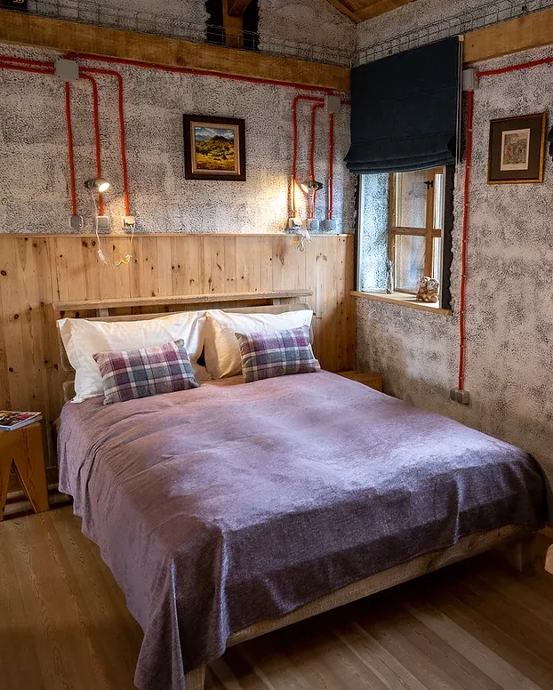 Уютна спалня с дървен интериор, съчетана с модерен дизайн. Стените са покрити с дървени панели до средата и текстуриран тапет отгоре. Леглото е оборудвано с комфортно двойно матрак, покрито с меко лилаво одеяло и подредени върху него карирани възглавници. Над леглото са монтирани декоративни стенни лампи. От едната страна на стаята се намира малък прозорец със завеса. Видими са и артистично подредени рамки с картини и декорации, придаващи допълнителен чар на стаята.