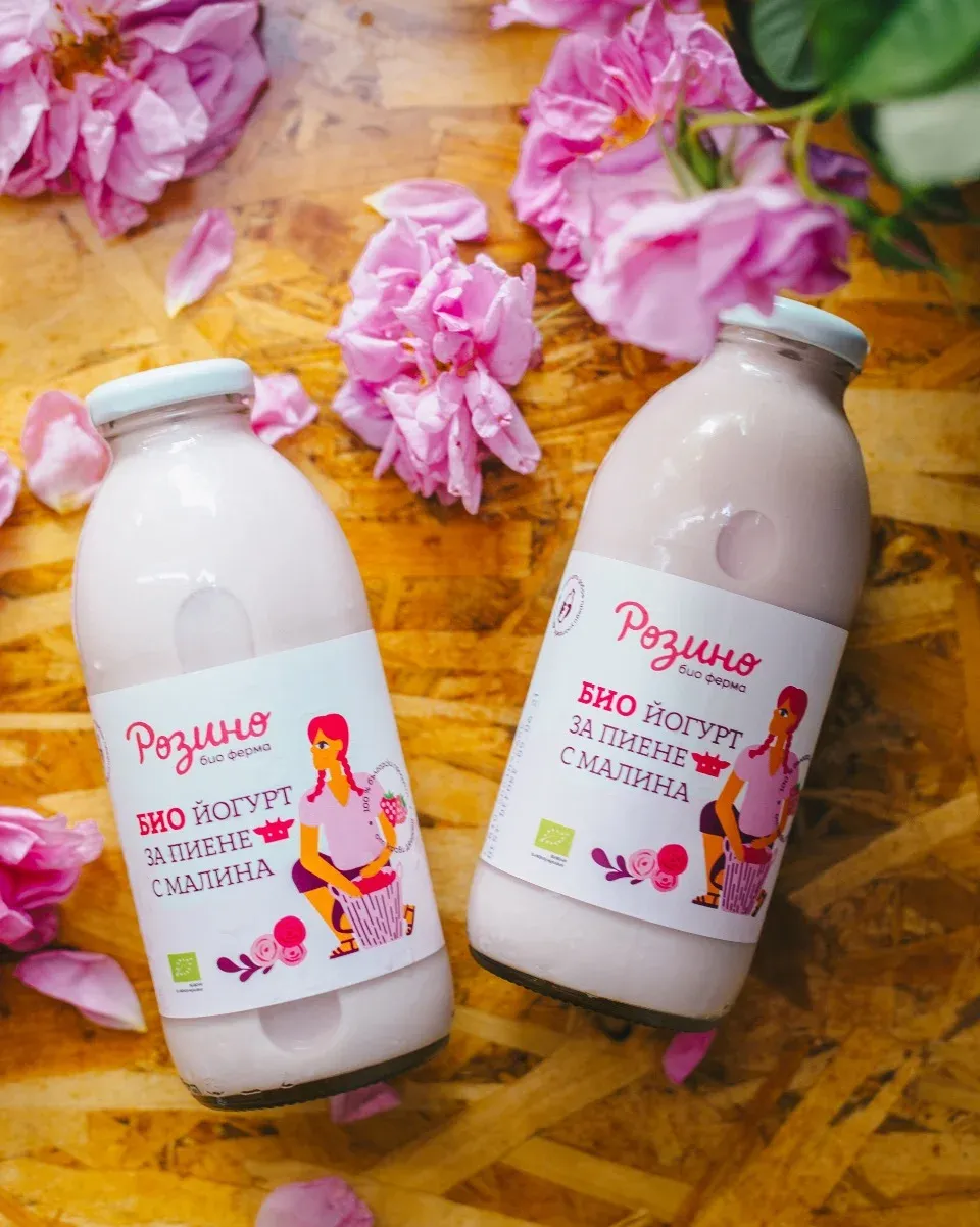 Две бутилки с био кефир на дървена маса, заобиколени от розови цветове. Бутилките са с етикети, на които е изобразена жена с кошница с цветя. На етикетите пише "Розино био ферма" и "Био йогурт за пиене с малина".