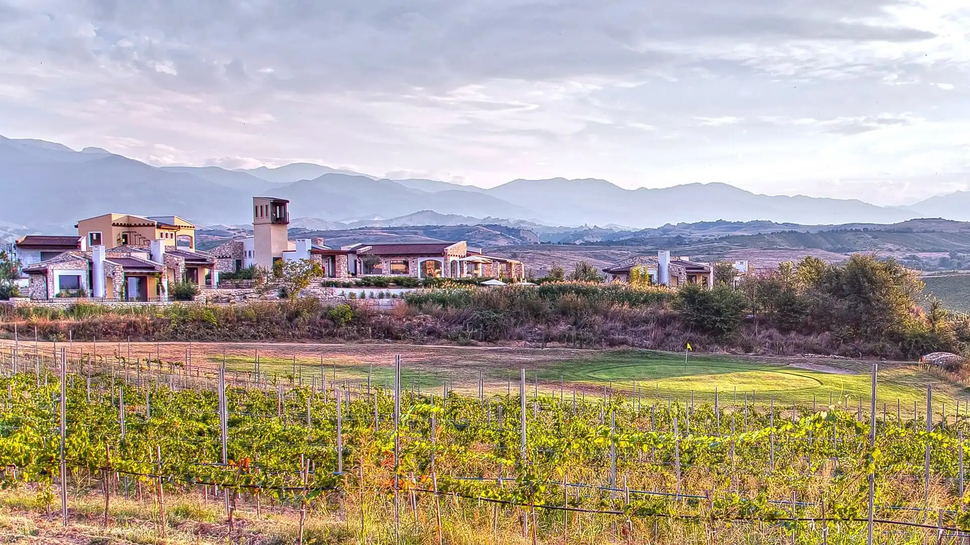 Панорамен изглед към винарска къща, разположена сред лозови масиви с планини на заден план при залез. Къщата е със съвременна архитектура, комбинираща камък и мазилка, и разполага с няколко структури с различни височини, включително висока кула.