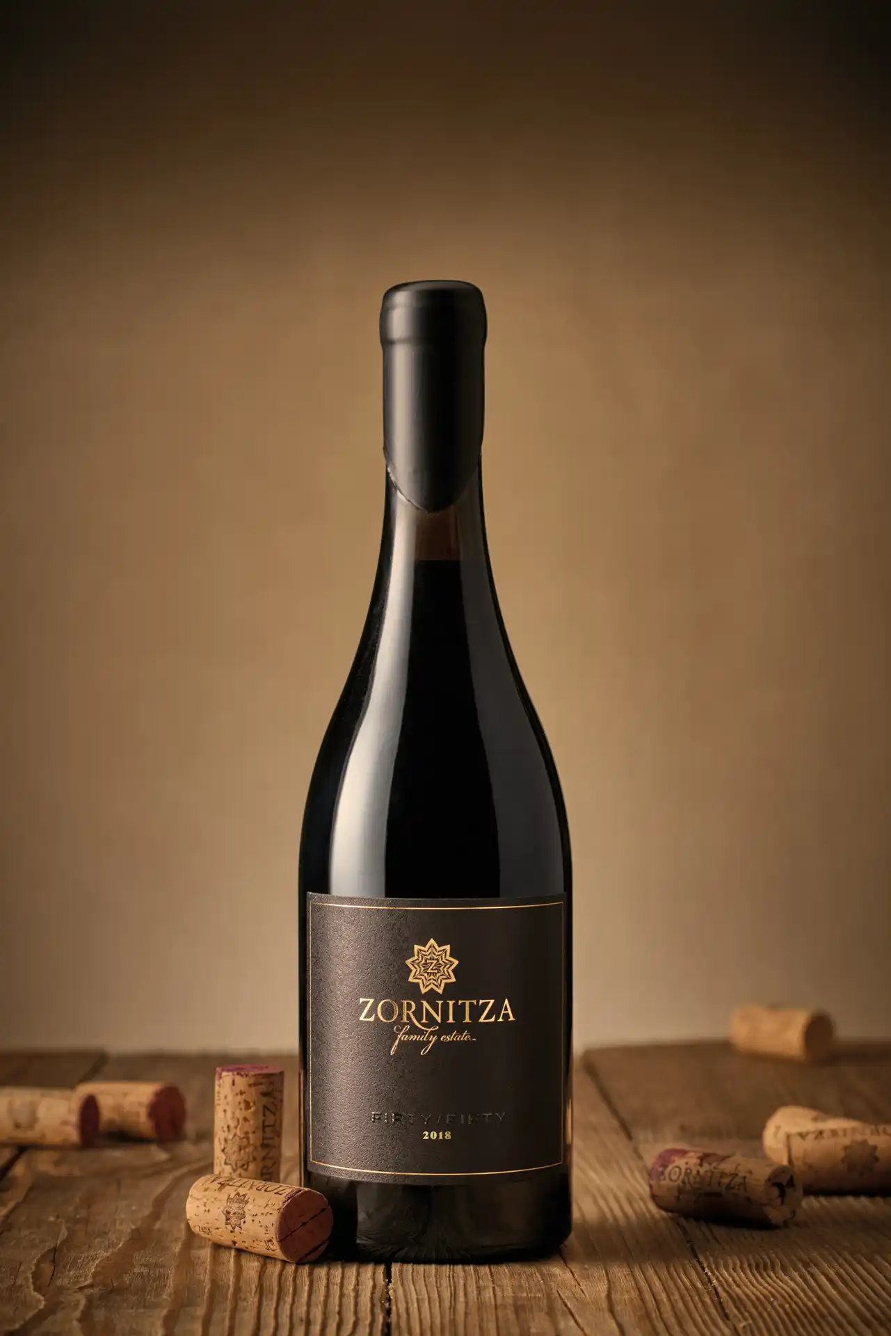 Бутилка вино "Зорница Family Estate Fifty/Fifty 2018" поставена изправено на дървена маса, с редица тапи разпилени около нея. Етикетът на бутилката е тъмносив със златни акценти, включително златна звезда в горната част, и представя логото на "Зорница". Фонът е меко осветен с нюанси на кафяво, което придава елегантен и изискан вид на цялата композиция.