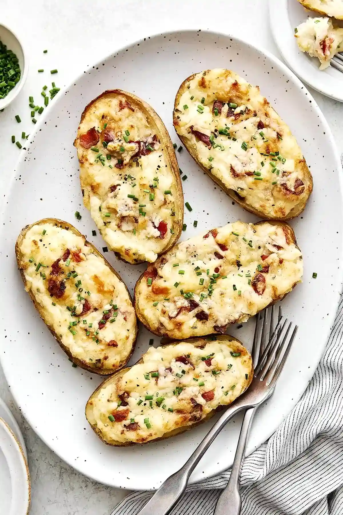 Печени картофи, пълнени с бекон и сирене, сервирани на бяла чиния. Всяко парче е посипано с нарязан зелен лук и черен пипер, придаващи апетитен вид на ястието. Допълнително, встрани са поставени вилица и салфетка, което подсказва за подредена трапеза.