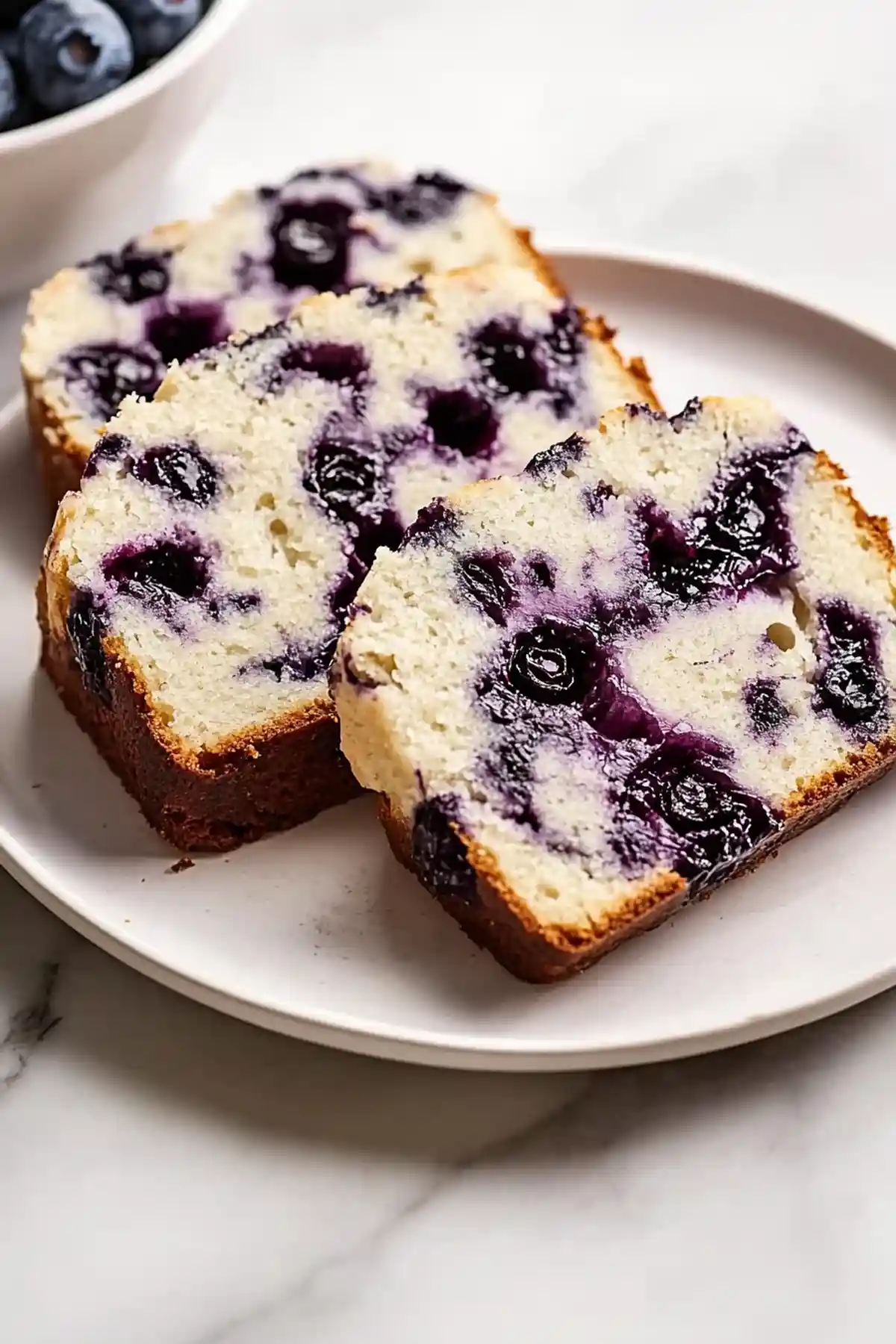 Две филии сладък кекс с боровинки на бяла чиния. Филиите имат златиста коричка и обилно количество боровинки, разпределени във вътрешността, заличаващи се до ярко лилаво. В задния план има бяла купа със свежи боровинки.