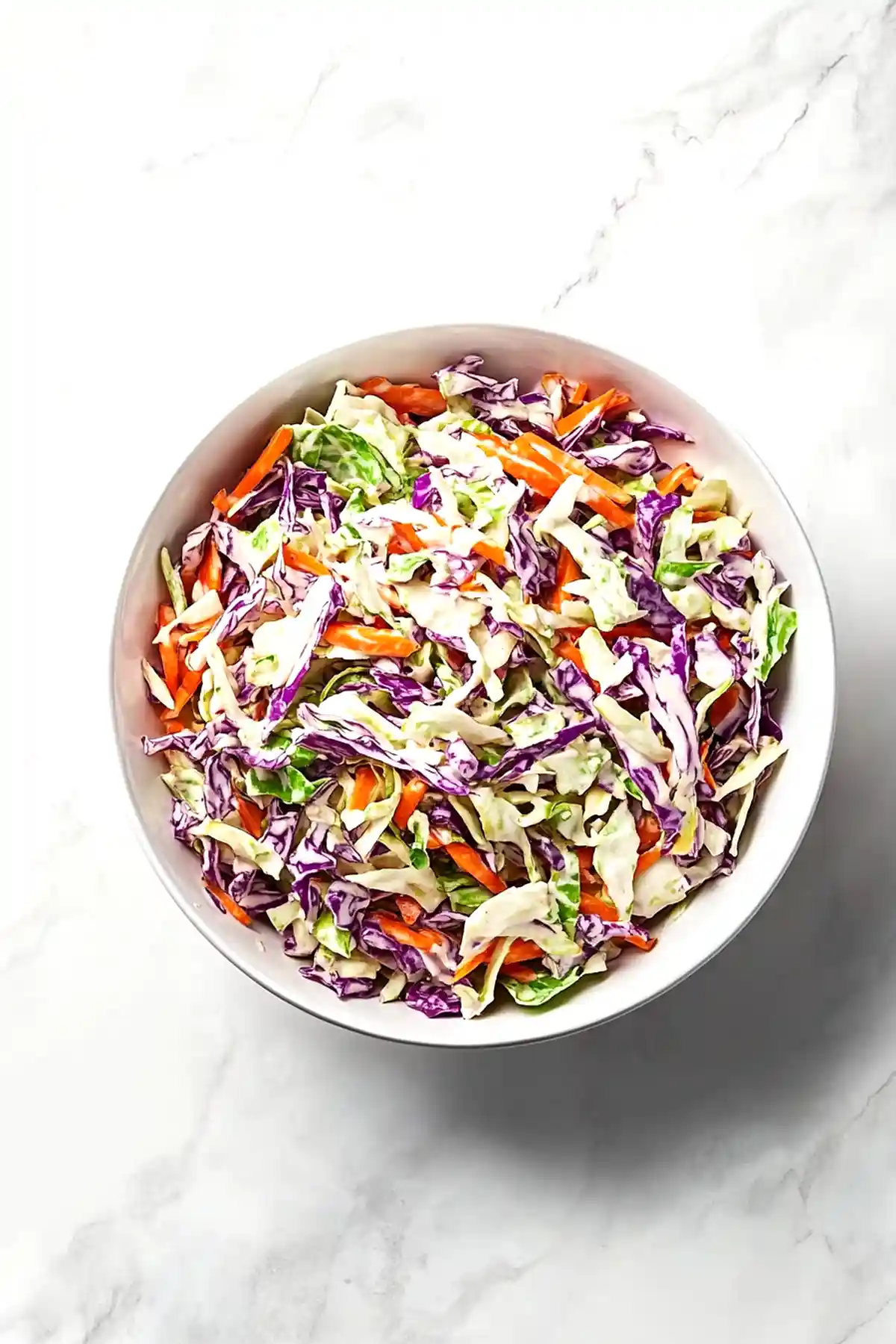 Салата Колсло от нарязани на ленти бяло и червено зеле, моркови и зелена салата, сервирана в бяла керамична купа върху светло мраморно бюро.