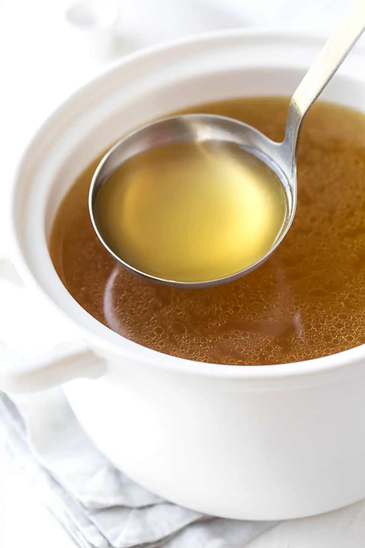 Бяла керамична купа с бульон, съдържащ жълто-златиста течност, с метална лъжица, която зачерпва от течността. Купата е на бял фон заедно с бяло кърпиче под нея.