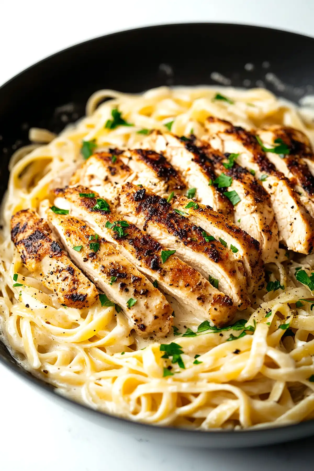 Черна керамична тиган с кремообразен фетучини паста, украсена с парченца нарязано пилешко месо отгоре, подправено с черен пипер и наръсено с пресен магданоз.