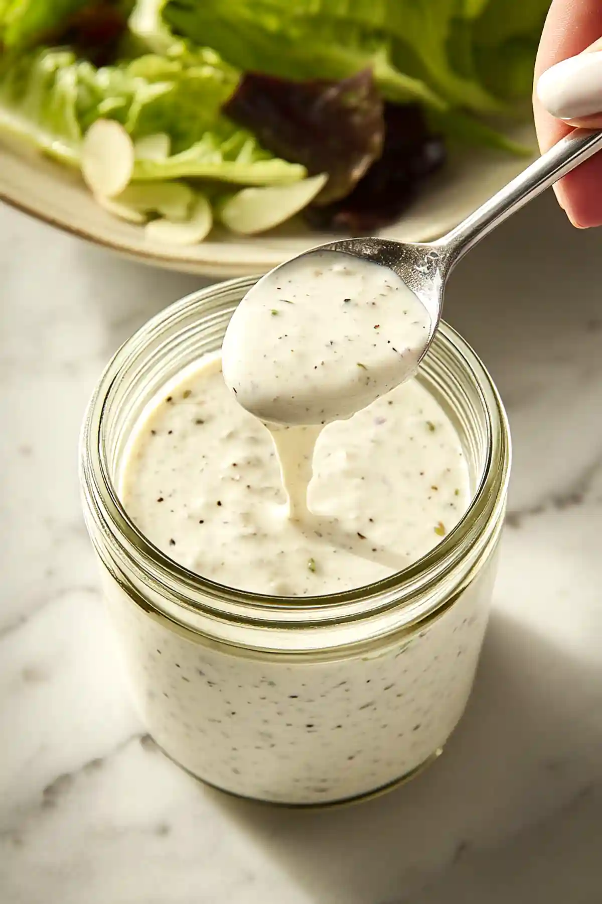 Лъжица с домашен сос ранч се изважда от стъклено бурканче на мраморна маса. На заден план се вижда чиния със салата от пресни зелени салатни листа и резени круша.