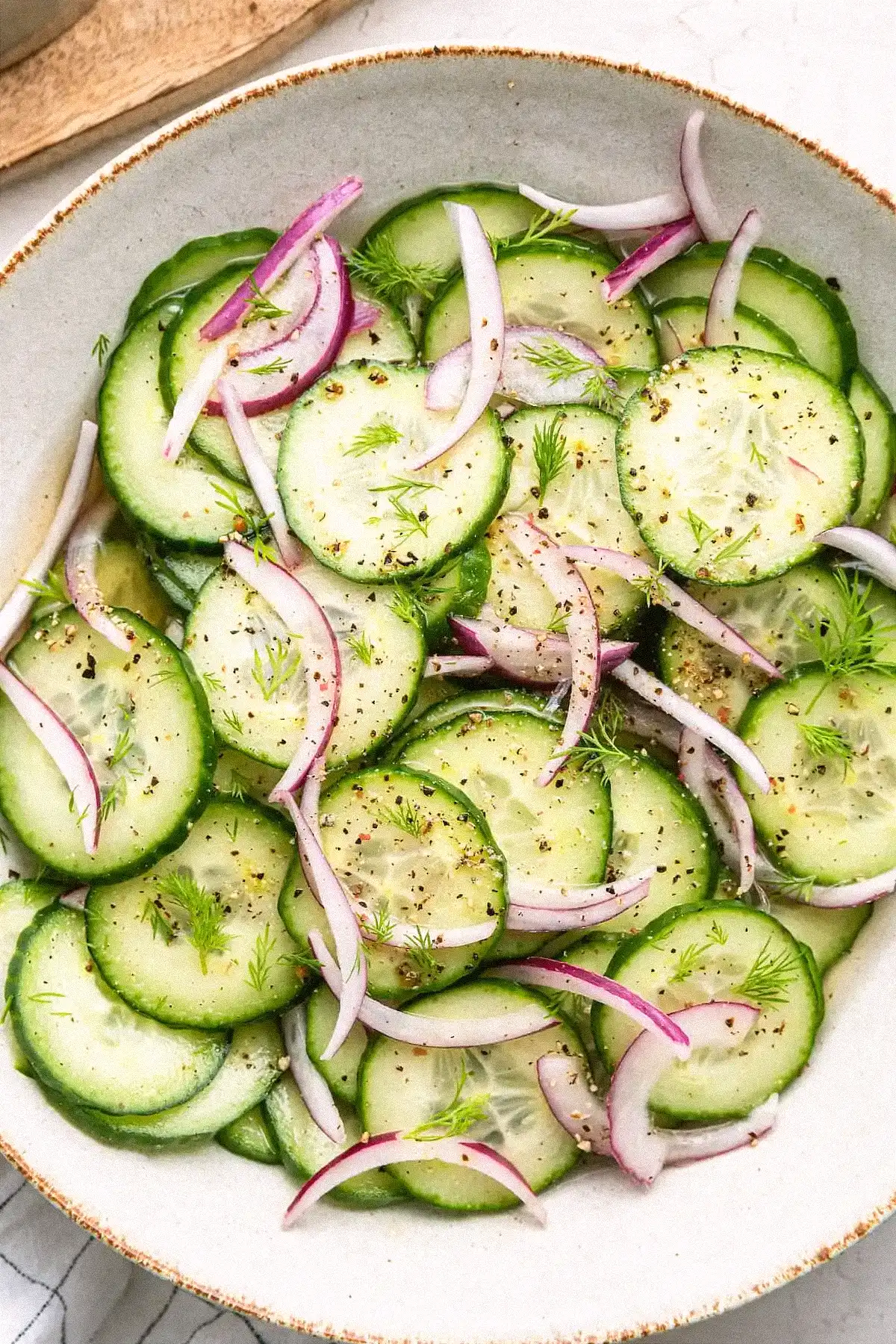 Свежа краставична салата с копър и червен лук, сервирана в дървена купа. Тънко нарязаните краставици са овкусени с винен оцет, зехтин, сол, черен пипер и пресен копър за ароматна и лека гарнитура.