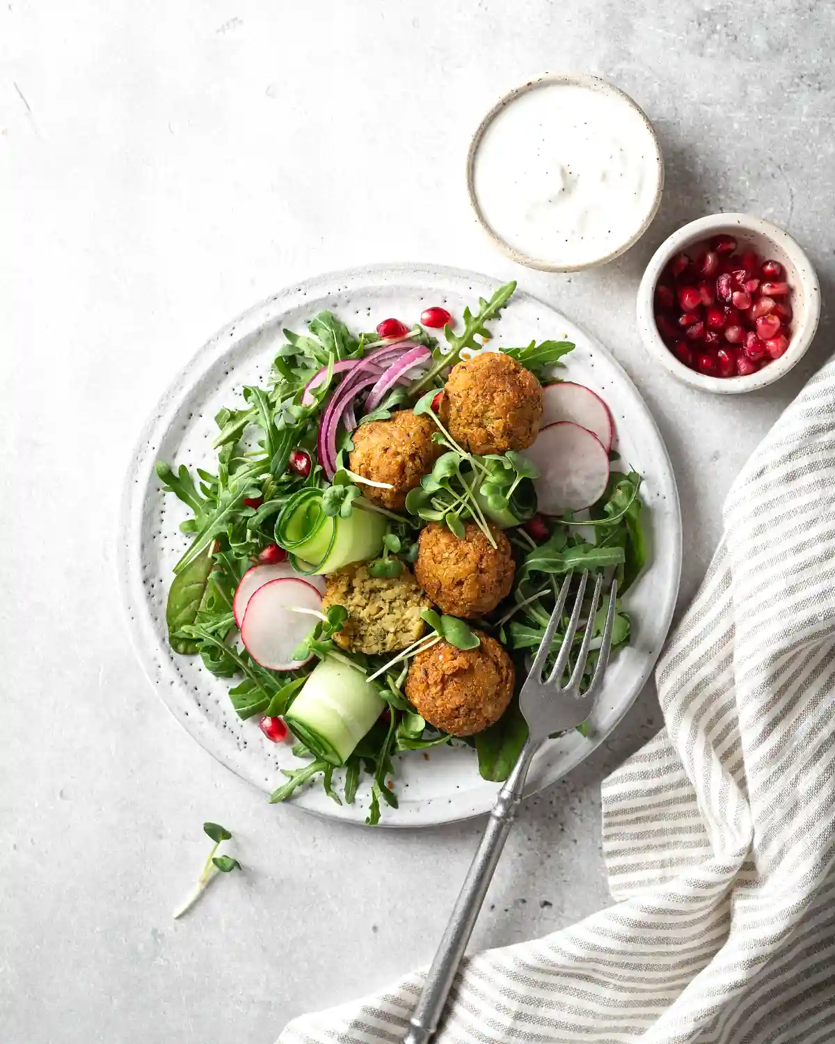 Бяла керамична чиния със зелена салата, където се открояват три златисти фалафели, нарязани краставици, репички, червен лук и гранатови зърна, заедно с малка купичка сос и покривка с бели и сиви райета отстрани.