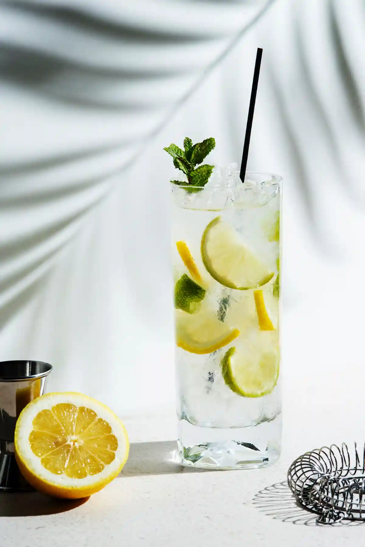 Висока стъклена чаша с освежаваща напитка, пълна с ледени кубчета, резени лимон и лайм, декорирана с листенца мента и черна сламка. На преден план на масата лежи половин лимон и метална смесителка за напитки, а на заден план има бяла драпирана завеса с меки сенки.