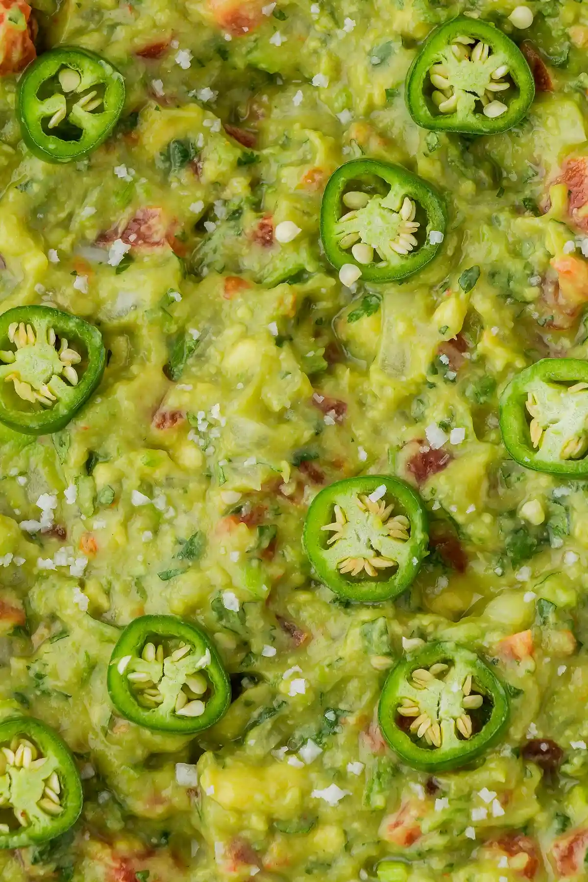 Крупен план на домашно приготвена гуакамоле с резени зелени люти чушки отгоре, наситено зелена със зърнаста текстура и видими парчета червен домат и зелени билки. Поръсена е със солените кристали сол.