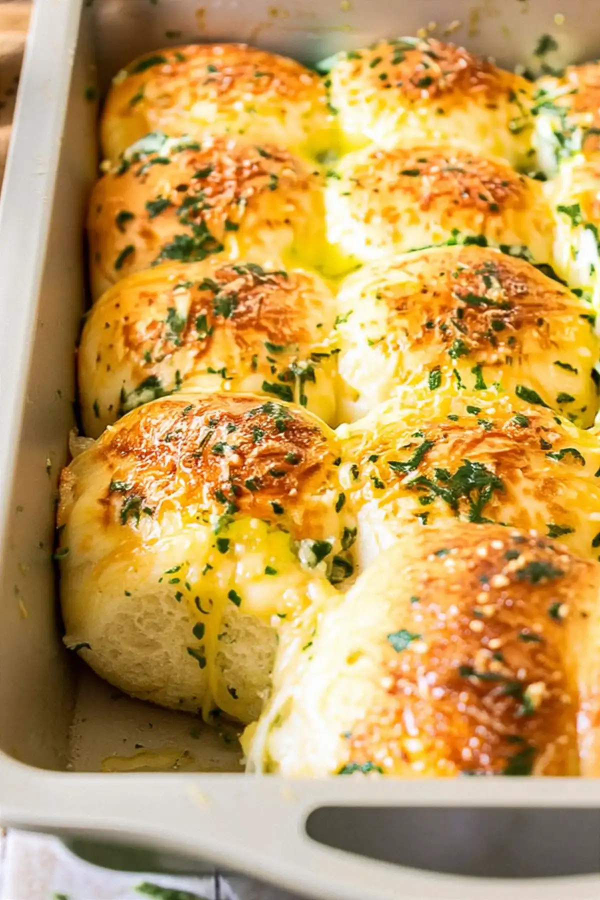 В керамичен съд са подредени поразяващо златисти и пухкави хлябчета, намазани с разтопено масло и поръсени обилно с наситнен магданоз. Хлябчетата блестят и изглеждат хрупкави отгоре, предразполагайки към апетитно изживяване.