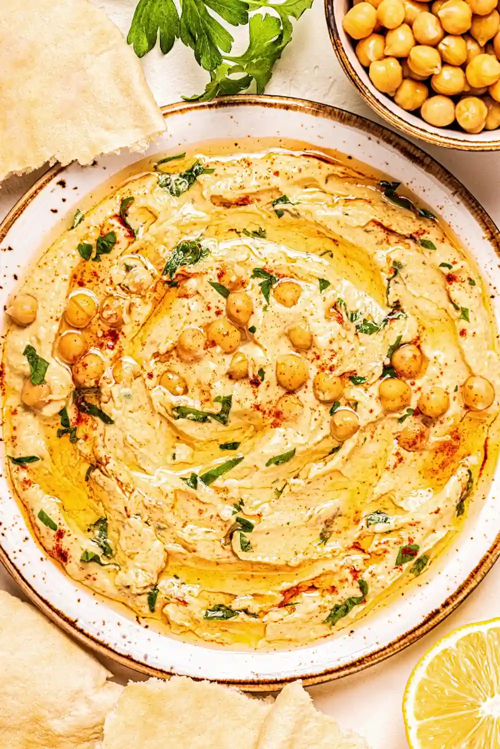 Порция от хумус в керамична чиния, украсена с нахутени резенчета, олио и подправки на финала. Отстрани се виждат резени лимон и купа с нахут, както и парчета ориенталски хляб. Цветовете са топли и апетитни, с акценти на зелено от билки и червено от паприка.