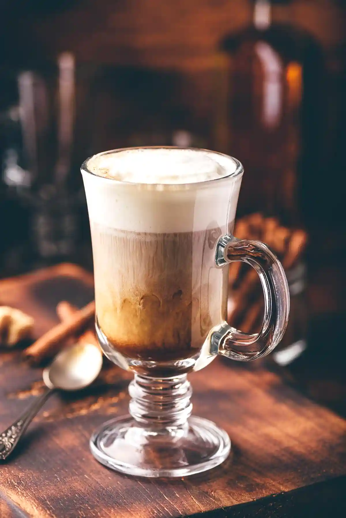 Чаша с Ирландско кафе на дървена маса, осветена с топла, мека светлина. Напитката е сервирана в прозрачна стъклена чаша с дръжка, показваща слоевете от кафе и обилна млечна пяна отгоре. До чашата има лъжица и фонът е тъмен с отблясъци на бутилки в задния план, което допринася за уютна и приветлива атмосфера.