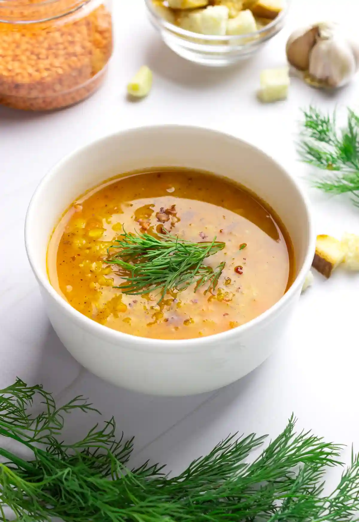 Бяла керамична купа с яркожълта лещена супа, поръсена с червен пипер и гарнирана с клонка копър. В задния план се виждат буркан с леща, чиния с кубчета хляб и скилидка чесън, заобиколени от свежи клонки копър.