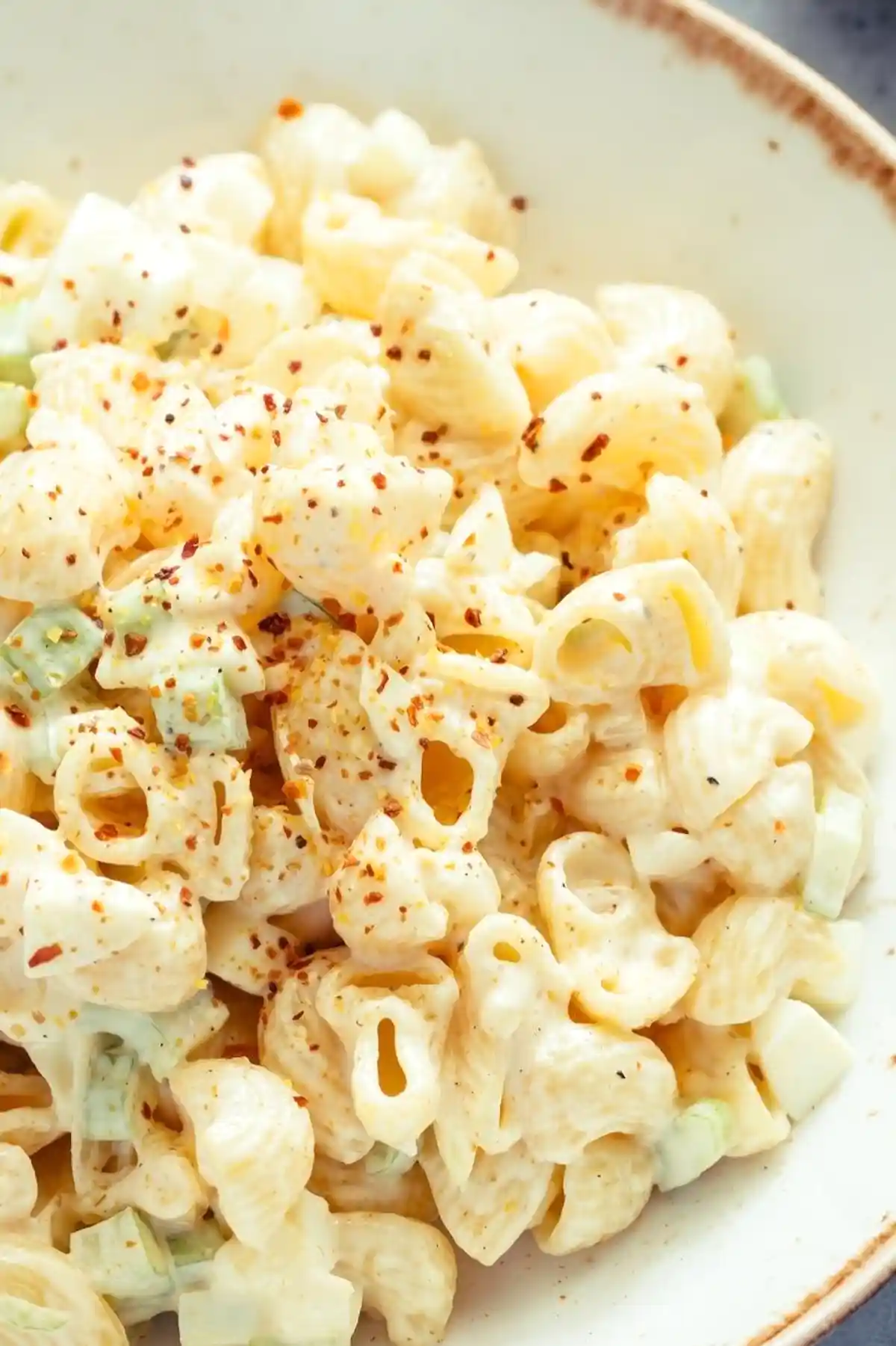 Порция салата от макарони в бяла керамична чиния, поръсена с червен пипер. Макароните са с форма на елица, облечени в кремообразен сос и с накълцан зелен лук.