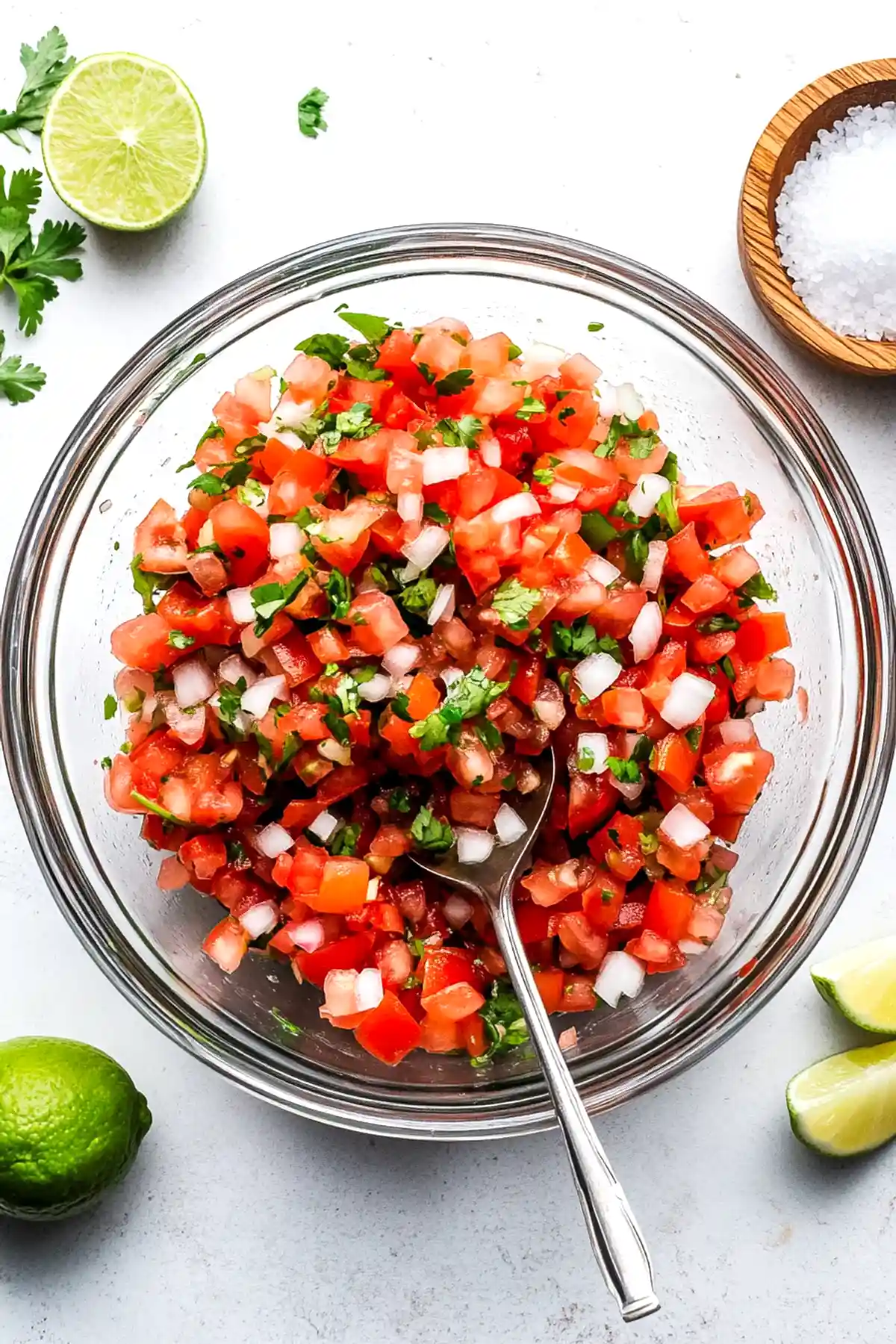 Прясно нарязана доматена салса Пико де Гайо в стъклена купа, подправена с лук, зеленина и кориандър, със сребриста лъжица вътре. Отстрани на масата има разположен половин лайм и дървен съд с морска сол.