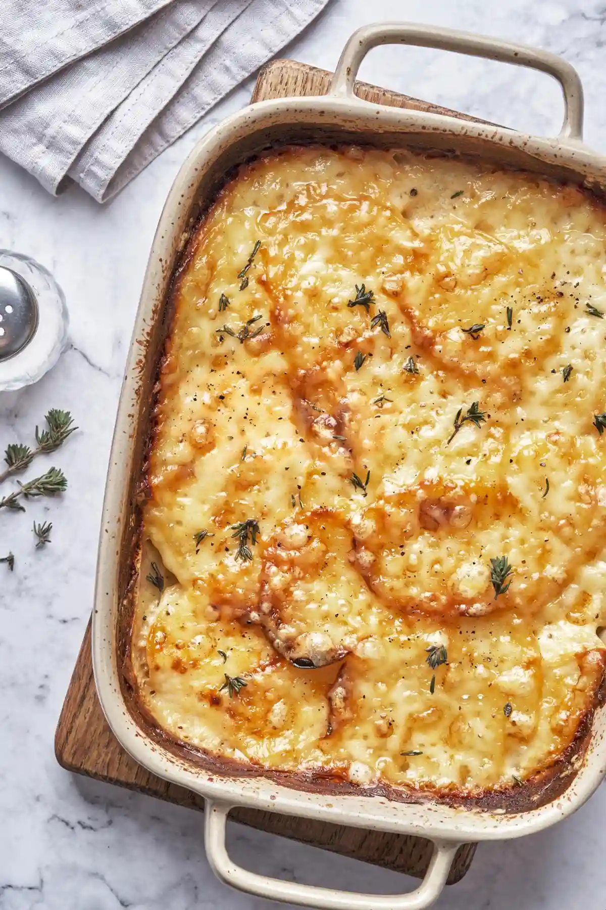 Ястие, изпечено в керамична тава, покрито със златисто кафяво разтопено сирене и поливка от масло. По повърхността са поръсени малки клонки пресен розмарин, добавяйки аромат и цвят. Фонът включва мраморна повърхност и сгъната сива салфетка, до тавата има дървена дъска.
