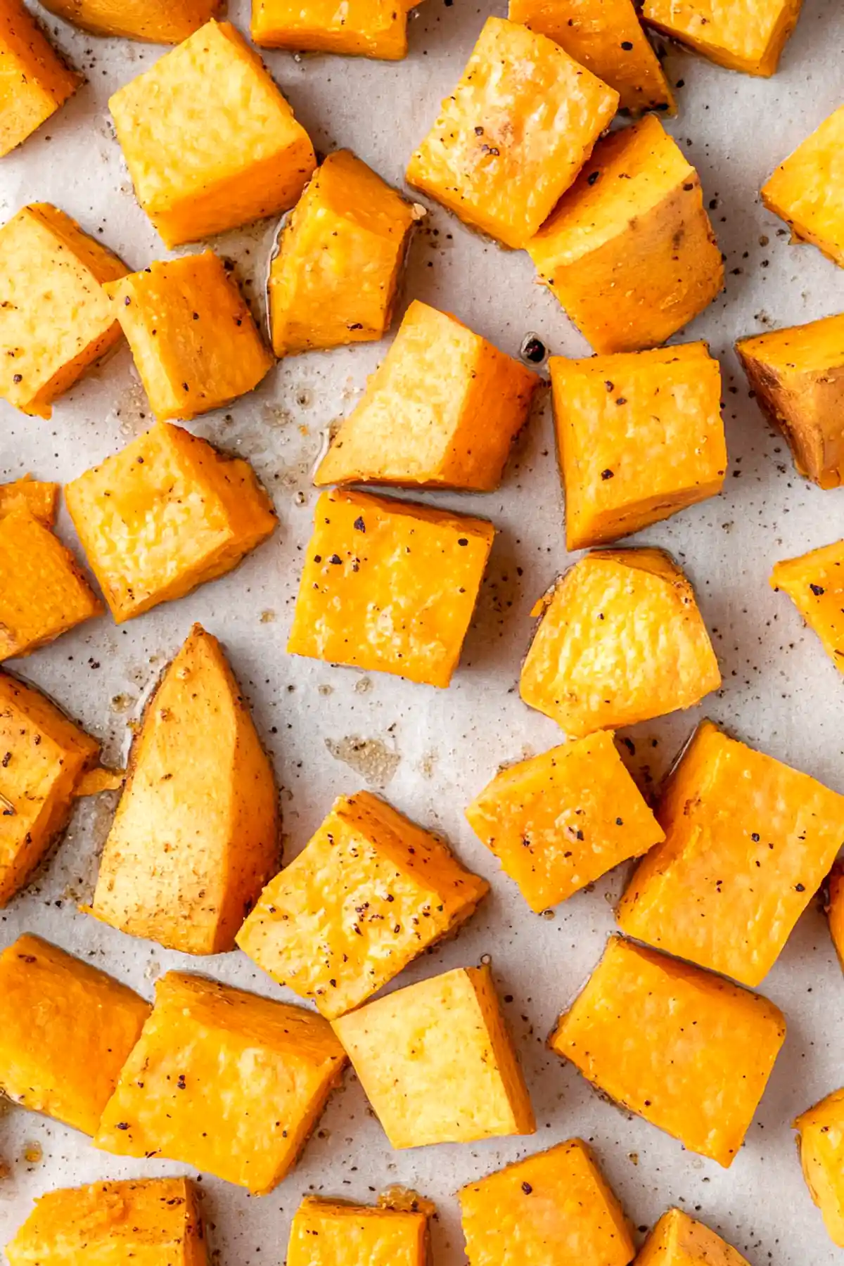 Печени парчета сладък картоф, подредени върху хартия за печене. Парчетата са неравномерно нарязани и захванати с черни подправки, което придава апетитен контраст на ярко оранжевия цвят на картофите.