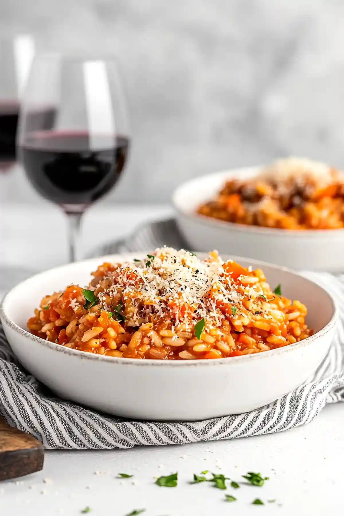 Бяла керамична чиния с порция Доматено ризото с червен сос, поръсена с пармезан и нарязани зелени билки, поставена на разноцветна кърпа. В задния план е частично фокусирана втора чиния със същото ястие и чаша червено вино.