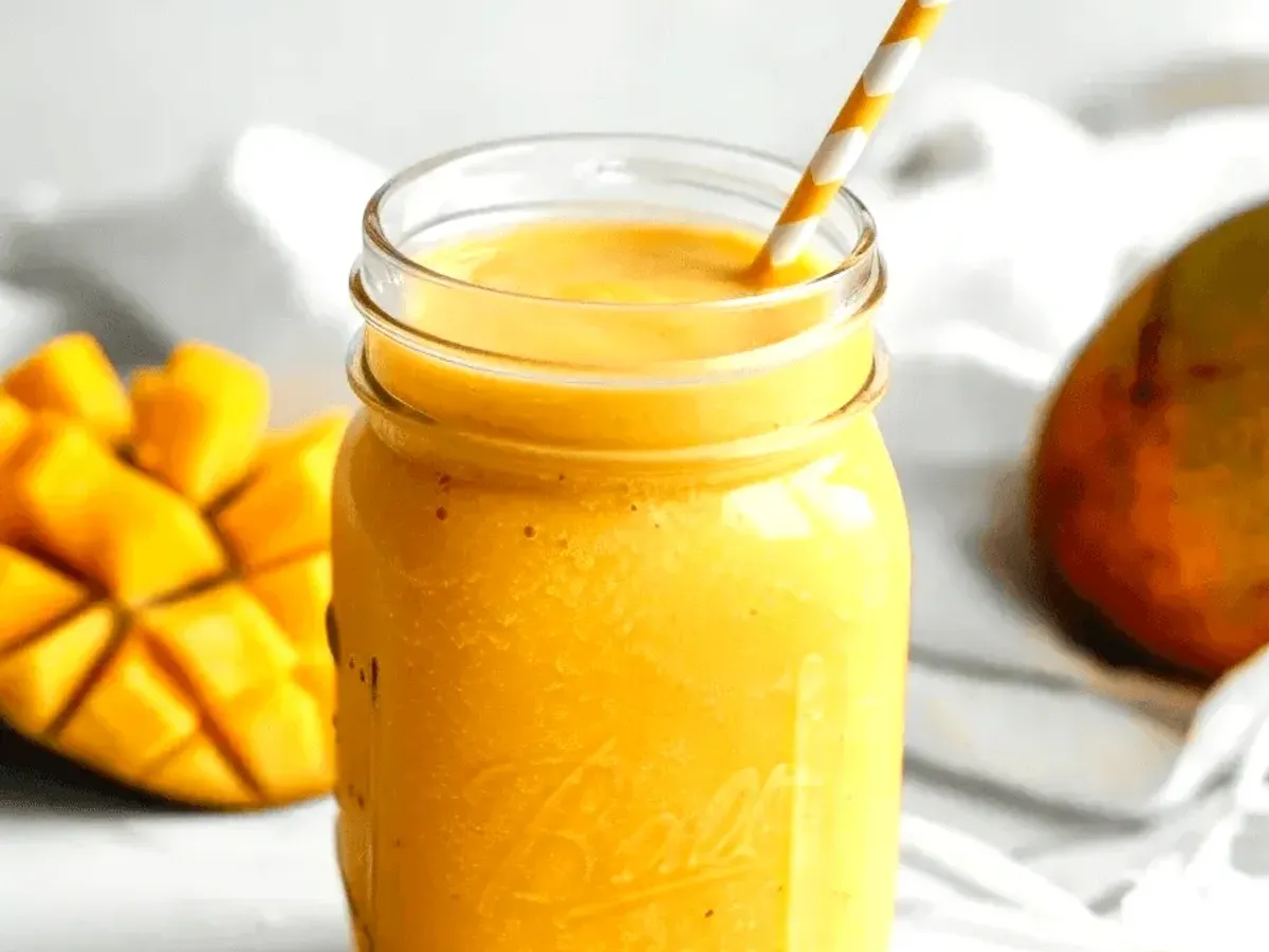 Стъклен буркан със златист манго смути, снабден с жълта сламка с бели точки. На заден план се намира частично нарязано манго и друго цяло манго на фокусиран бял фон.