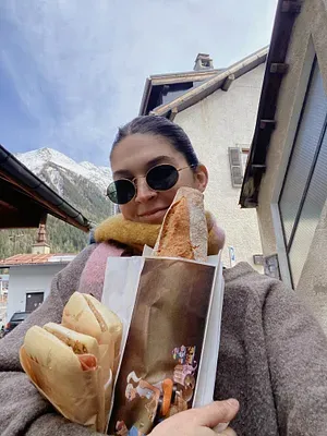 A woman wearing round sunglasses and a scarf tucked into a grey coat, holds up a large bag filled with assorted breads, including a baguette sticking out playfully. She is outdoors with a backdrop of a quaint building and snow-dusted mountains under a clear sky.
