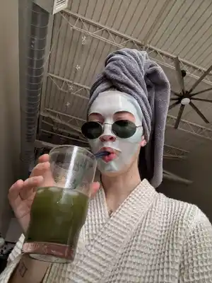 A person in a spa-like setting, wearing sunglasses, a facial mask, and a towel wrapped around their head. They are sipping a green smoothie through a straw and wearing a white robe. The background shows a ceiling with exposed beams and a large fan.